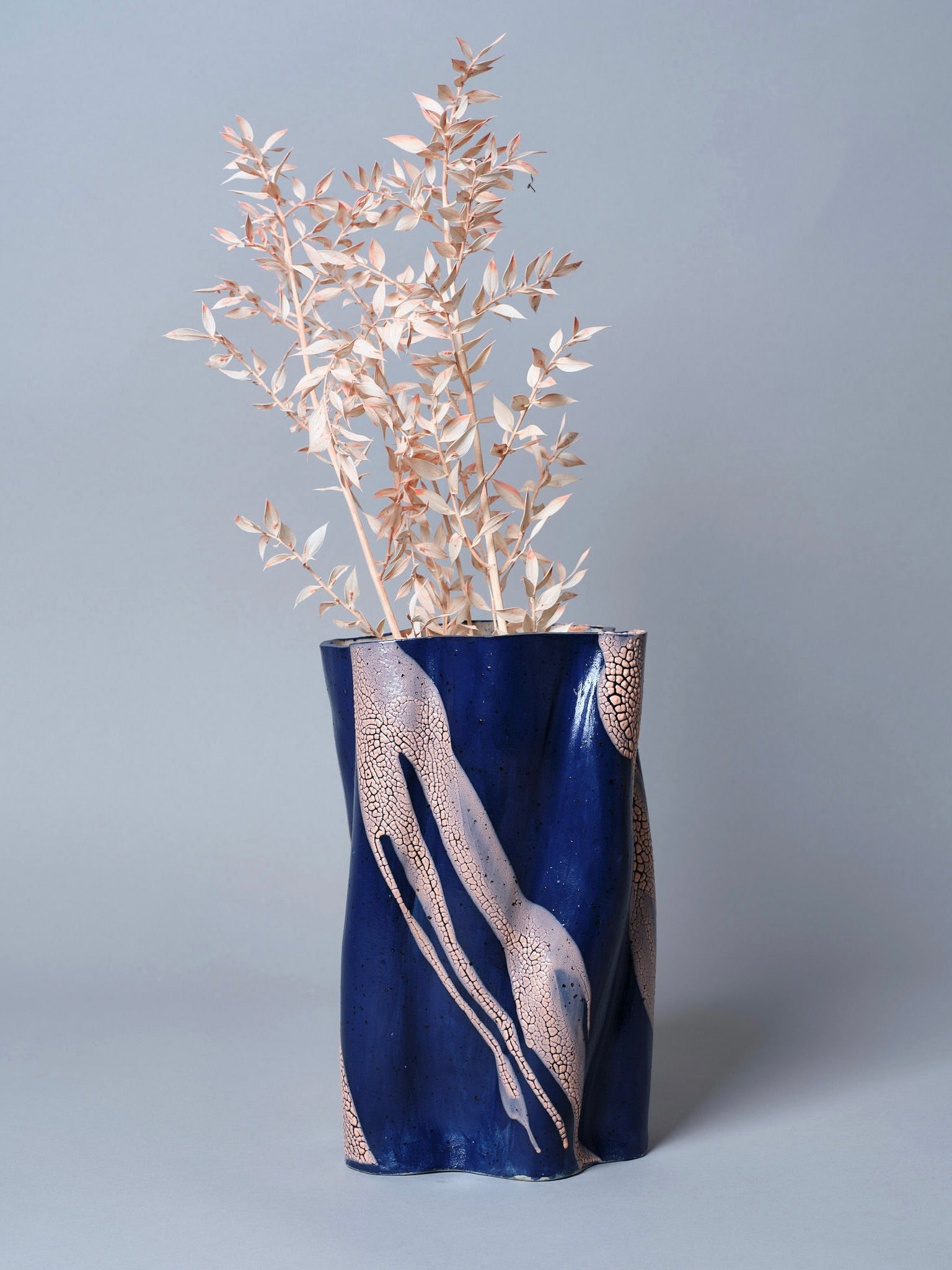 The Swirly Vase Blue and Peach by Astrid Öhman, with intricate patterns and filled with dried branches of light-colored leaves, stands elegantly against a plain gray background, showcasing a blend of traditional craftsmanship and modern elegance.