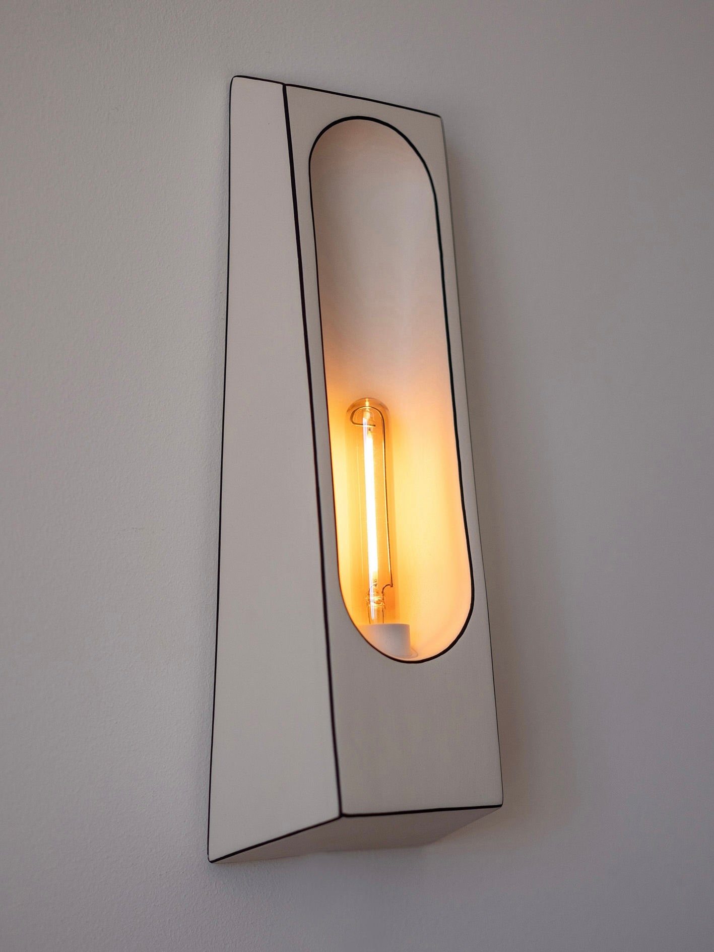 The Alcove Facette Wall Lamp by Violaine d'Harcourt is a modern, wall-mounted fixture with a hand-finished rectangular design. Its narrow oval opening showcases a vintage-style incandescent bulb, providing an accent light with a warm glow against the plain background.