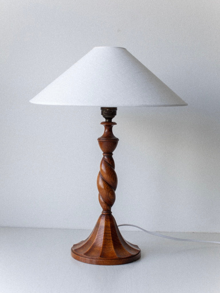 The Early 1900's Table Lamp by Out For Lunch, featuring a twisted wooden base and a white conical lampshade, exudes vintage decor charm. Placed against a plain, light-colored background, its power cord is slightly visible at the base. The design hints at an Art Deco influence that effortlessly elevates any space.
