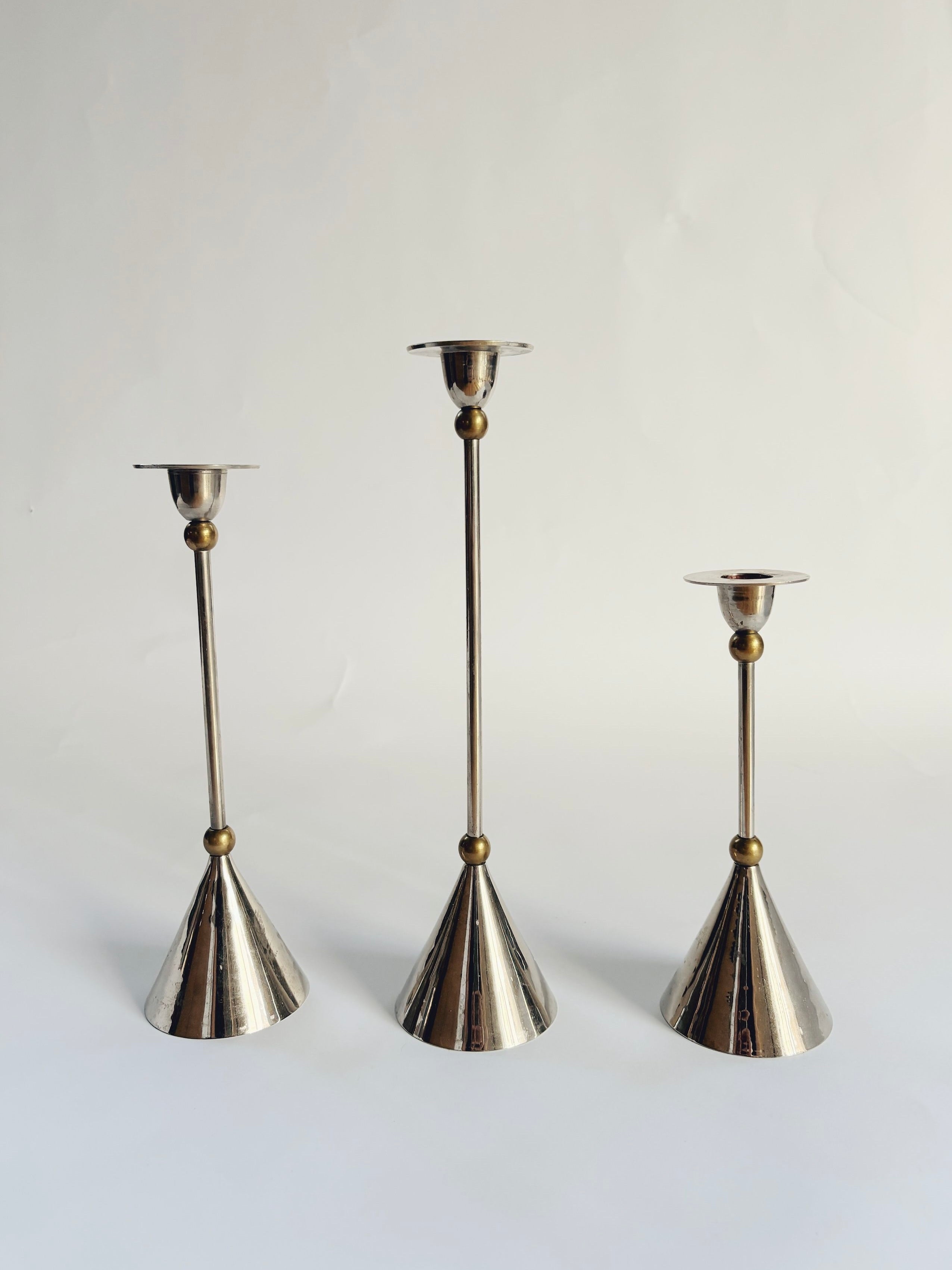 Amidst a plain backdrop, the Huerto Set of 3 Postmodern Candle Holders, with their varying heights, conical bases, and spherical accents, offer a discreet homage to 1980s design. The chrome and brass finishes capture light splendidly, embodying a minimalist yet sophisticated Memphis style aesthetic.