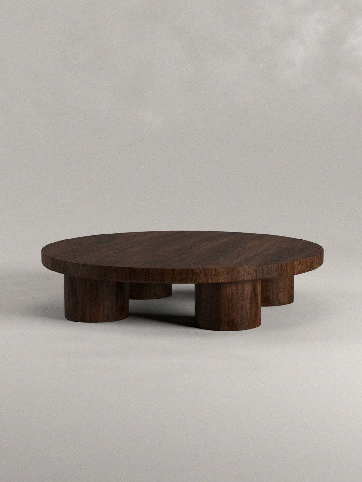 The Siete Studio Totem Coffee Table is handcrafted from solid walnut, featuring a round, low design with thick cylindrical legs. Its minimalist style accentuates the natural grain and rich color of the wood against a neutral backdrop.