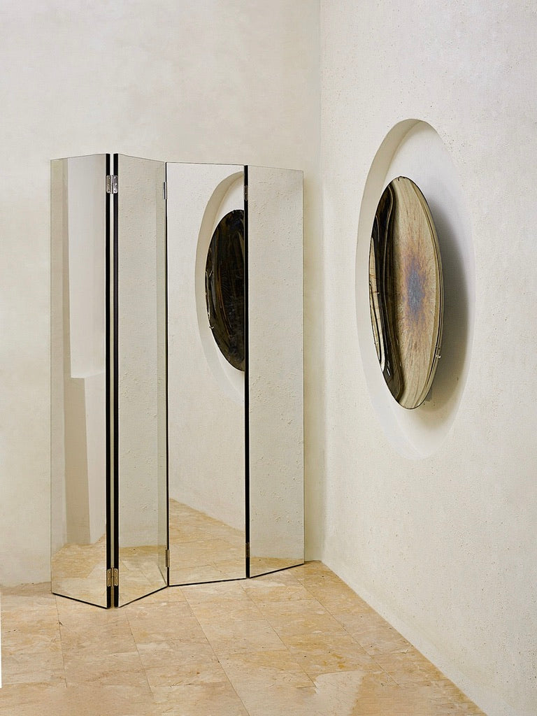 In a corner on the beige stone floor stands 'The Paravan' Mirror Folding Screen by HOMA. Next to it, a round abstract art piece is mounted on a circular recess in the cream-colored wall. The room boasts minimalist and modern elegance with unique, made-to-order charm.