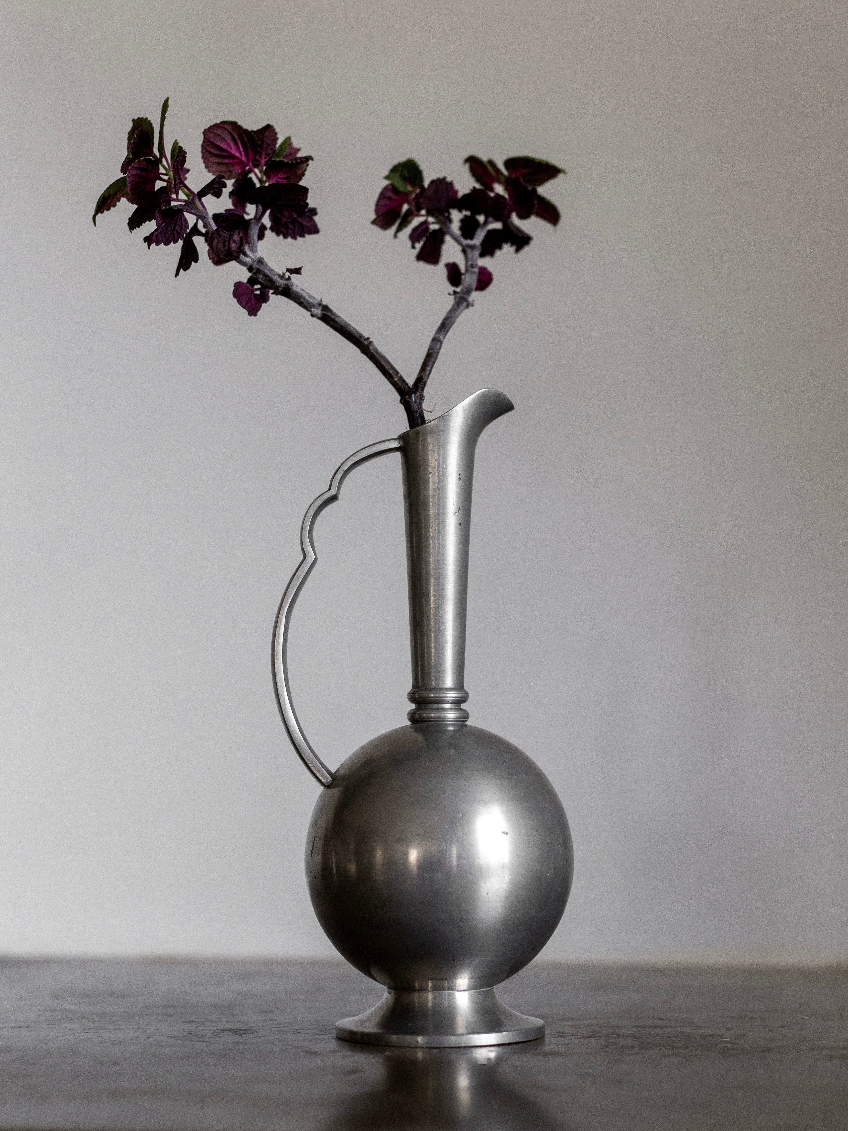 A minimalist still life photograph features an Art Deco Metal Vase from the 1930s by Spigel, showcasing an elegantly curved handle and a round base. The vase holds two dark foliage stems, standing against a plain gray background.