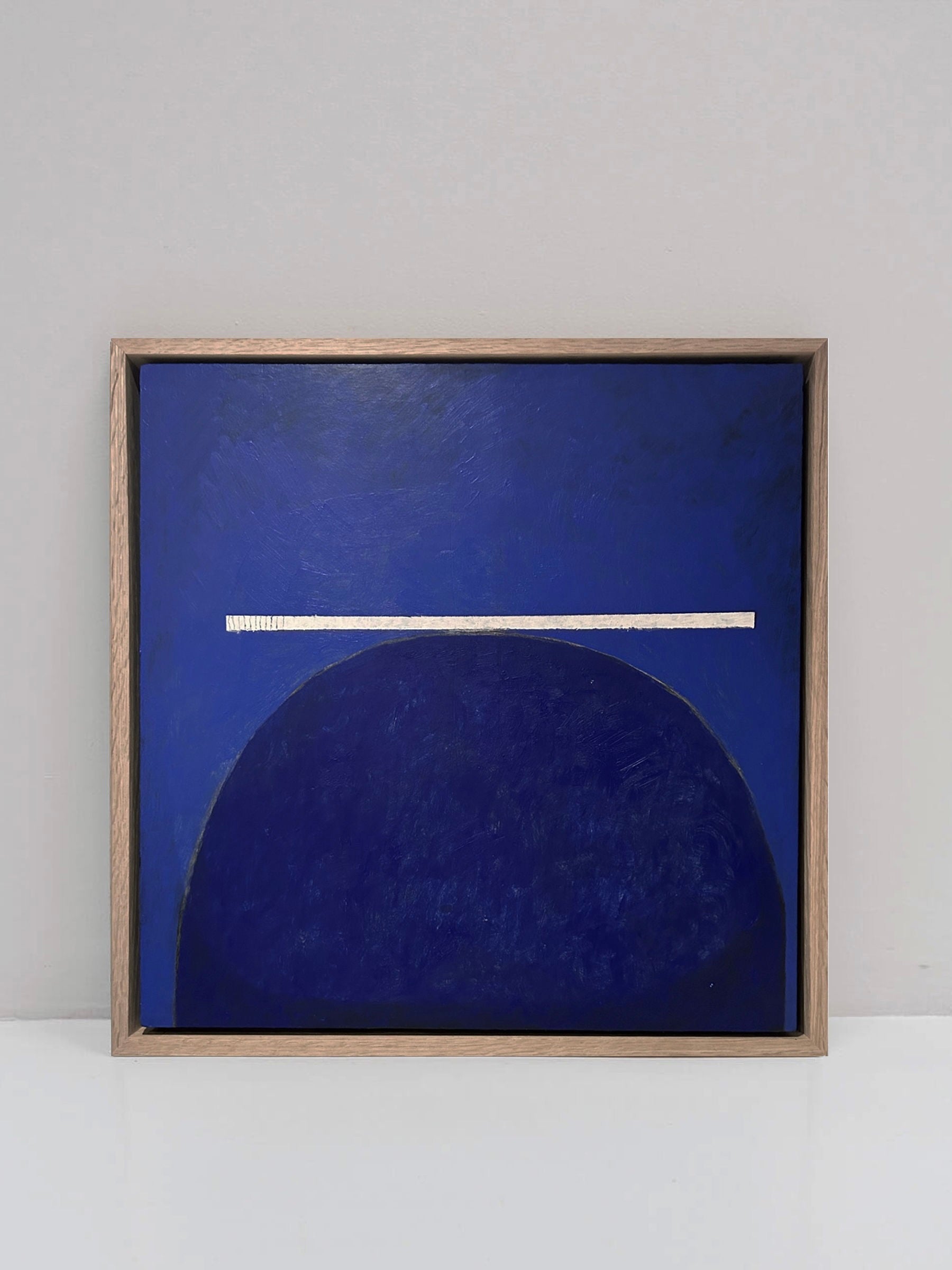 Introducing the "Balance" Painting by Lara Voce, a minimalist abstract masterpiece crafted with acrylic on a cradled wooden panel. It comes elegantly displayed in a wooden frame, featuring a deep blue semi-circle topped by a thin white line against a plain gray backdrop. Enjoy complimentary shipping on this captivating artwork.