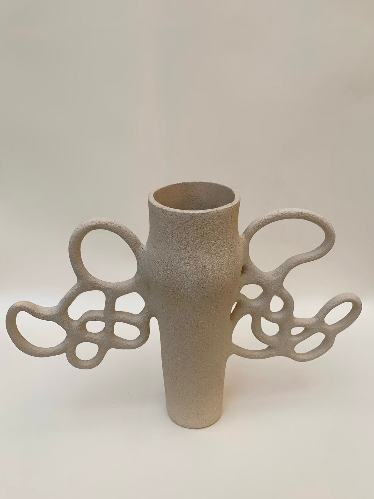 Part of the Louana Roudot Ceramic collection, the Vase Liens n°1 is a beige, abstract sculptural piece featuring circular and organic cutouts that extend like wings. Crafted from white chamotte stoneware, its matte texture elegantly stands against a neutral background.