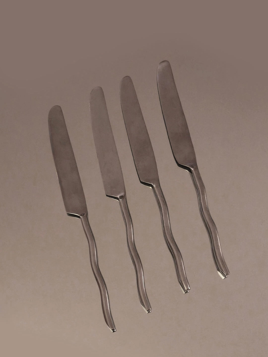 A set of four Wave Knives from Les Objoies, featuring silver-colored handles, is arranged parallel on a brown surface.