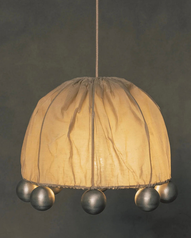 The "ALTER" Pendant Light by Kaytar has a hanging design with a beeswax-coated linen lampshade, featuring a gathered pattern and decorative silver balls around the bottom edge, casting a warm glow against dark backgrounds.