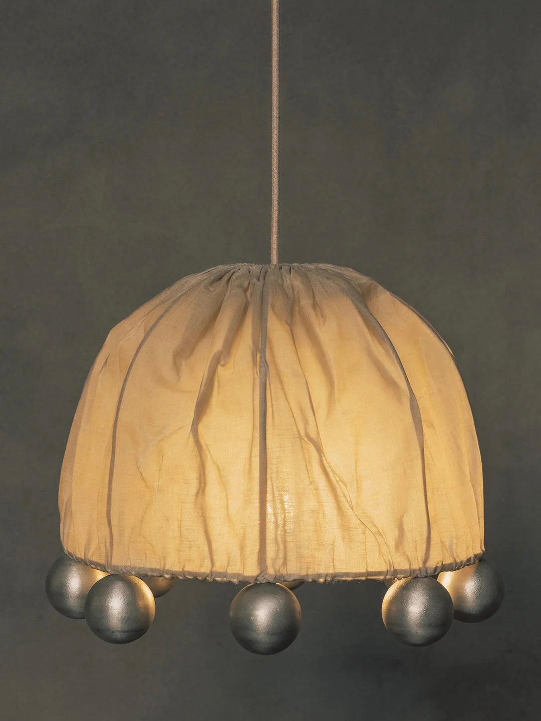 The "ALTER" Pendant Light by Kaytar has a hanging design with a beeswax-coated linen lampshade, featuring a gathered pattern and decorative silver balls around the bottom edge, casting a warm glow against dark backgrounds.