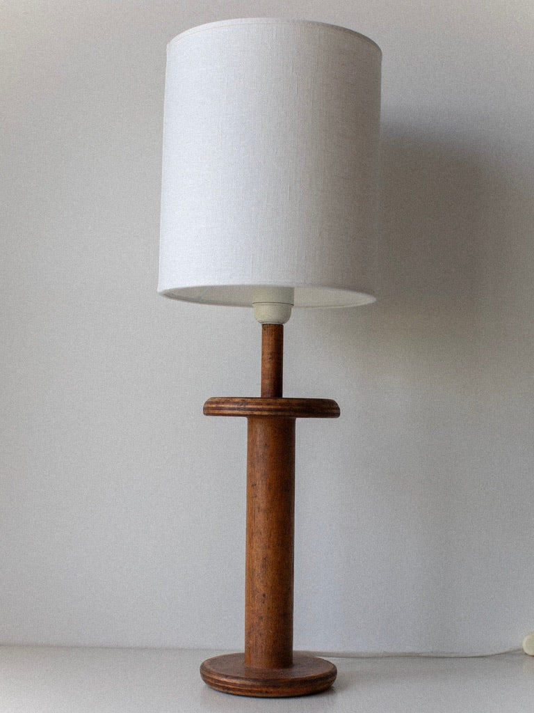 A Midcentury Table Lamp from Out For Lunch, featuring a wooden cylindrical base paired with a white drum lampshade, is placed against a plain white background. The lamp's simple, minimalist design exudes timeless elegance.