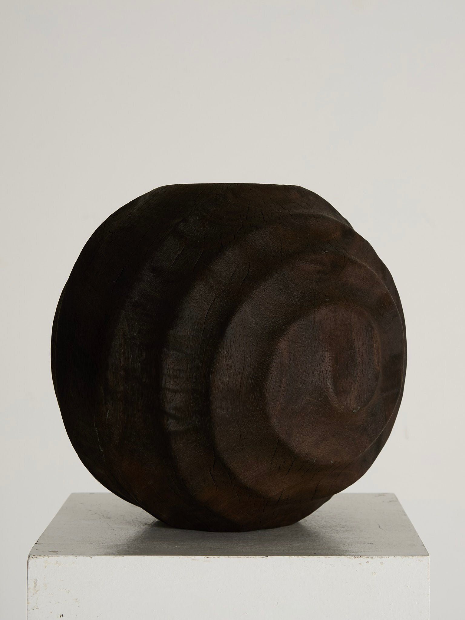 The Hardwood Sculptured Vessel by Ancien et Jolie, a textured dark wood piece from 1970s Britain with intricate circular patterns, sits elegantly on a white pedestal against a plain light background.