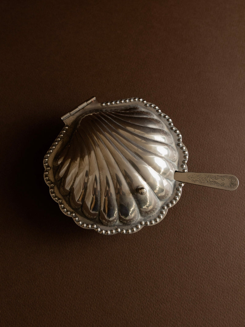 A closed Silver-plated Butter/Caviar Dish by Bottega Jacobs rests on a dark brown surface, featuring a scalloped shell-shaped design with a small ornate spoon. This vintage piece, crafted in Italy during the 1950s, boasts a hinged lid and a textured finish that reflect its elegant charm and timeless allure.