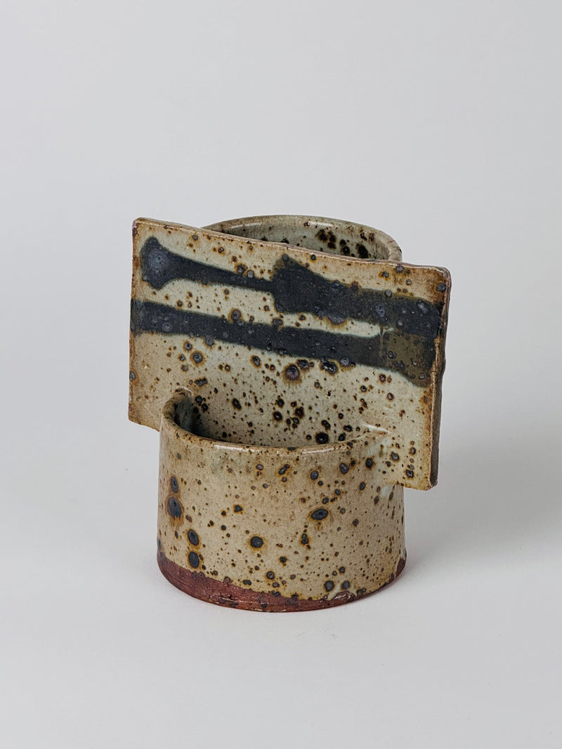 The Geometric Vase by Gustave Tiffoche, a Figuresdesign creation, is a ceramic sculpture with a cylindrical base topped by a rectangular piece. The speckled beige glaze has spots of brown and black, and the rectangular segment resembles an abstract double vase adorned with black line patterns.