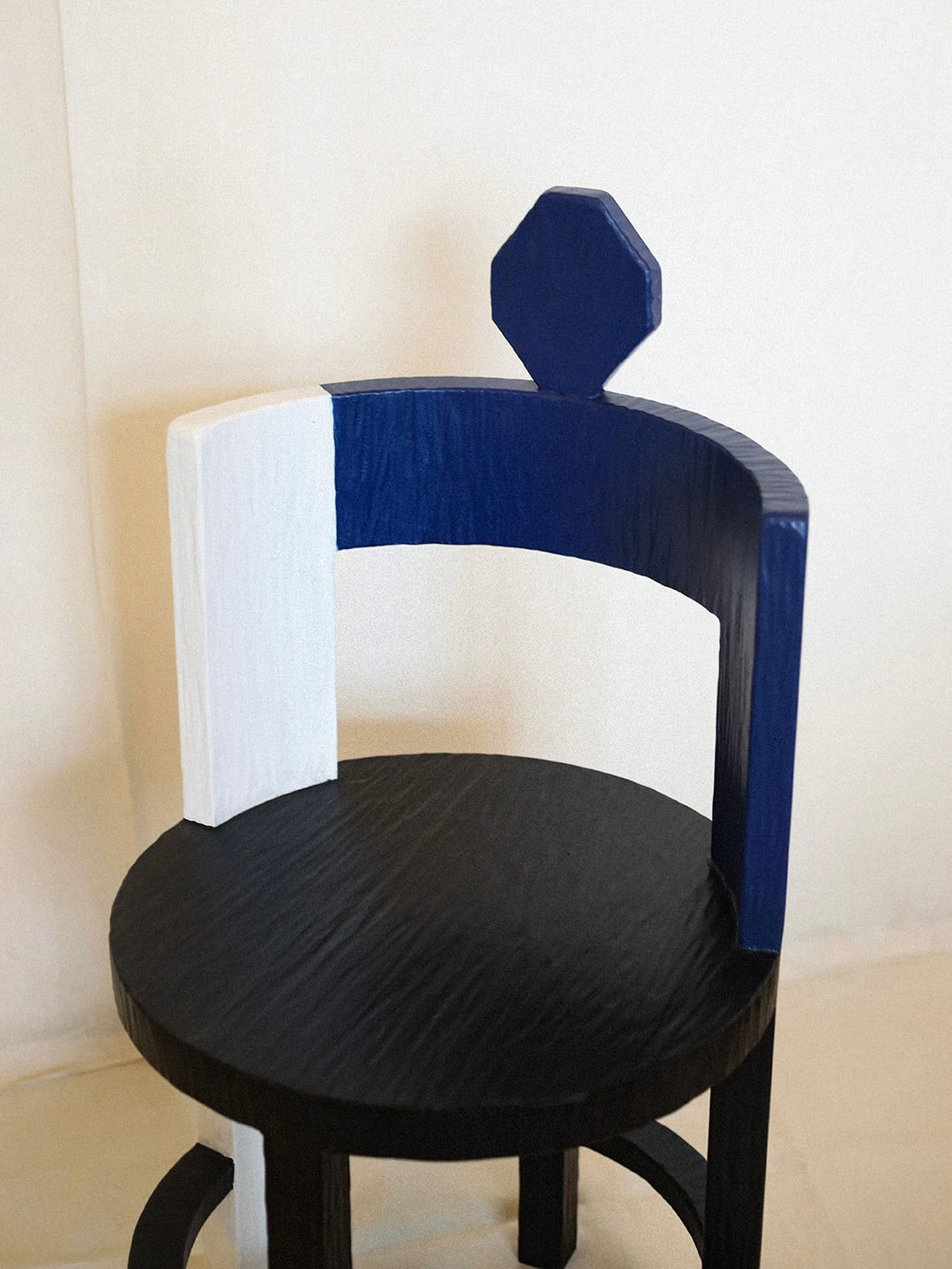 The Sculptural Chair I in Primary Colors by Rooms Studio showcases artistry and sustainability with a rounded black seat, three reclaimed oak legs, and a geometric semi-circle backrest in white and dark blue, topped with a hexagonal shape. It stands against a plain background.