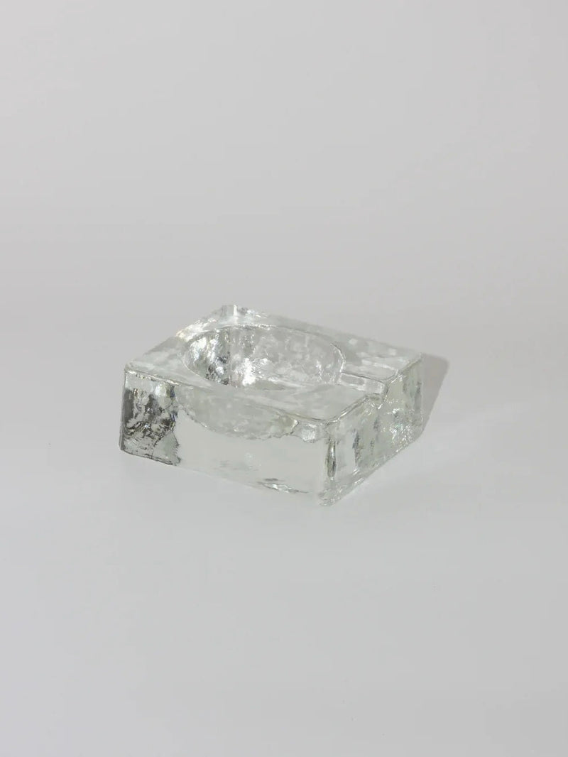 A rectangular, clear glass Brutalist Ashtray by Boga Avante Shop, featuring a central depression, rests on a plain light gray surface. The vintage ashtray's design is modern and minimalist, seamlessly blending Brutalist style with geometric form and smooth edges.