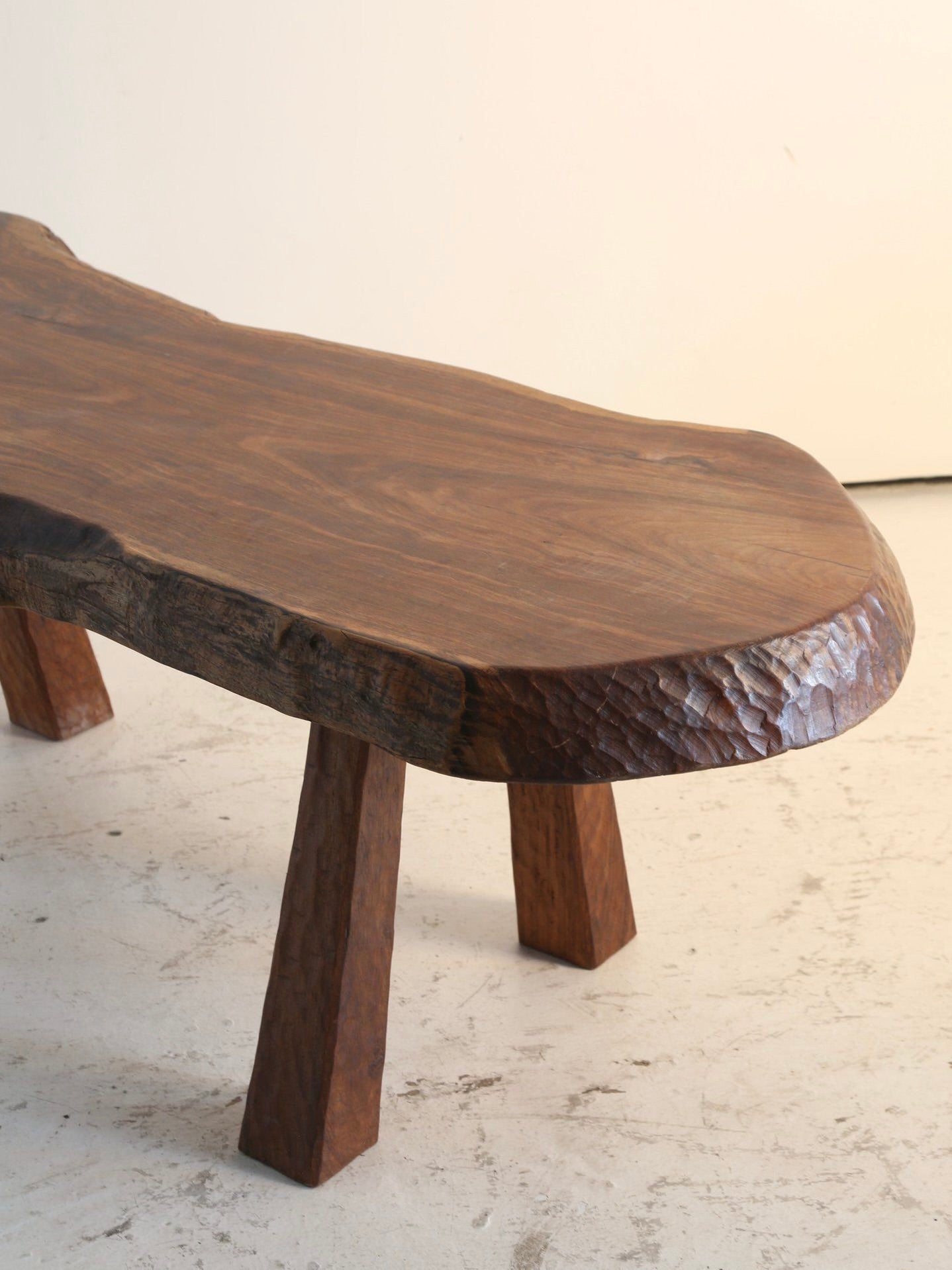 The 'French Folk' Coffee Table by Ancien et Jolie features a rustic waney edge, irregular top, and three sturdy angled legs. Adze carved details highlight its rich dark grain and smooth surface, echoing the timeless charm of France's 1970s design with textured carvings on edges and legs.