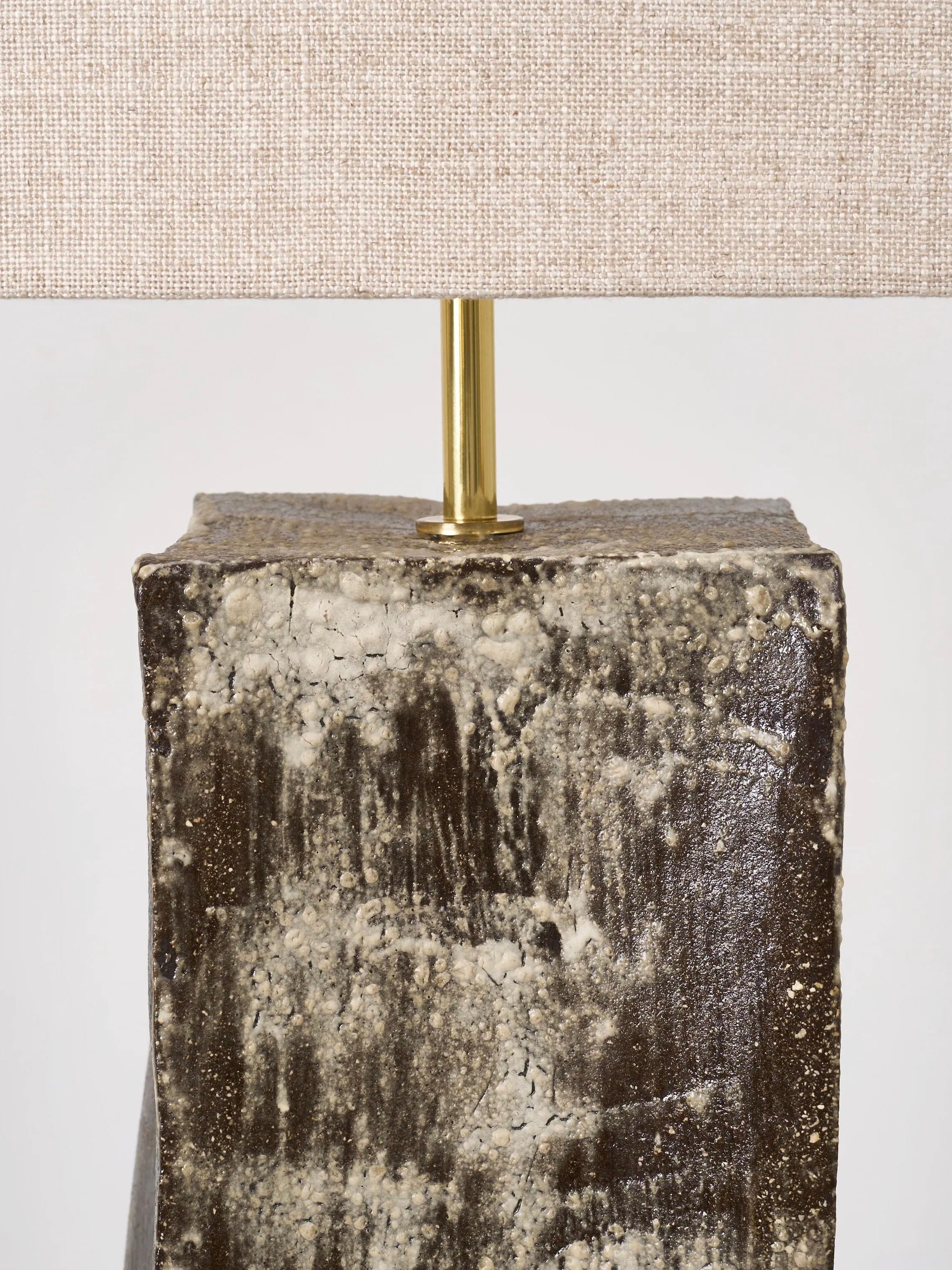 Close-up of Project 213A's Rectangular Ceramic Table Lamp, showcasing a handmade rectangular base with a rough, textured surface in dark brown and speckled gray tones. A gold-colored rod supports the cream fabric lampshade above this artisanal ceramic light.