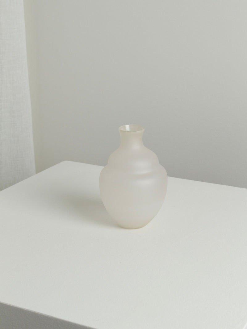 The Small Vase in Moon by Verre d'Onge, with its frosted glass finish, round base, and narrow neck, is placed on a white surface against a minimalist background. The soft, moon-like diffused light highlights the vase's subtle texture and translucent quality, evoking the tranquil ambiance of an evening in Montreal.