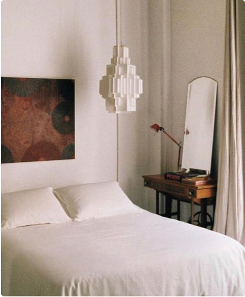 Minimalist bedroom with a white bed, modern geometric pendant light, and a small wooden desk with a mirror in a serene atmosphere.