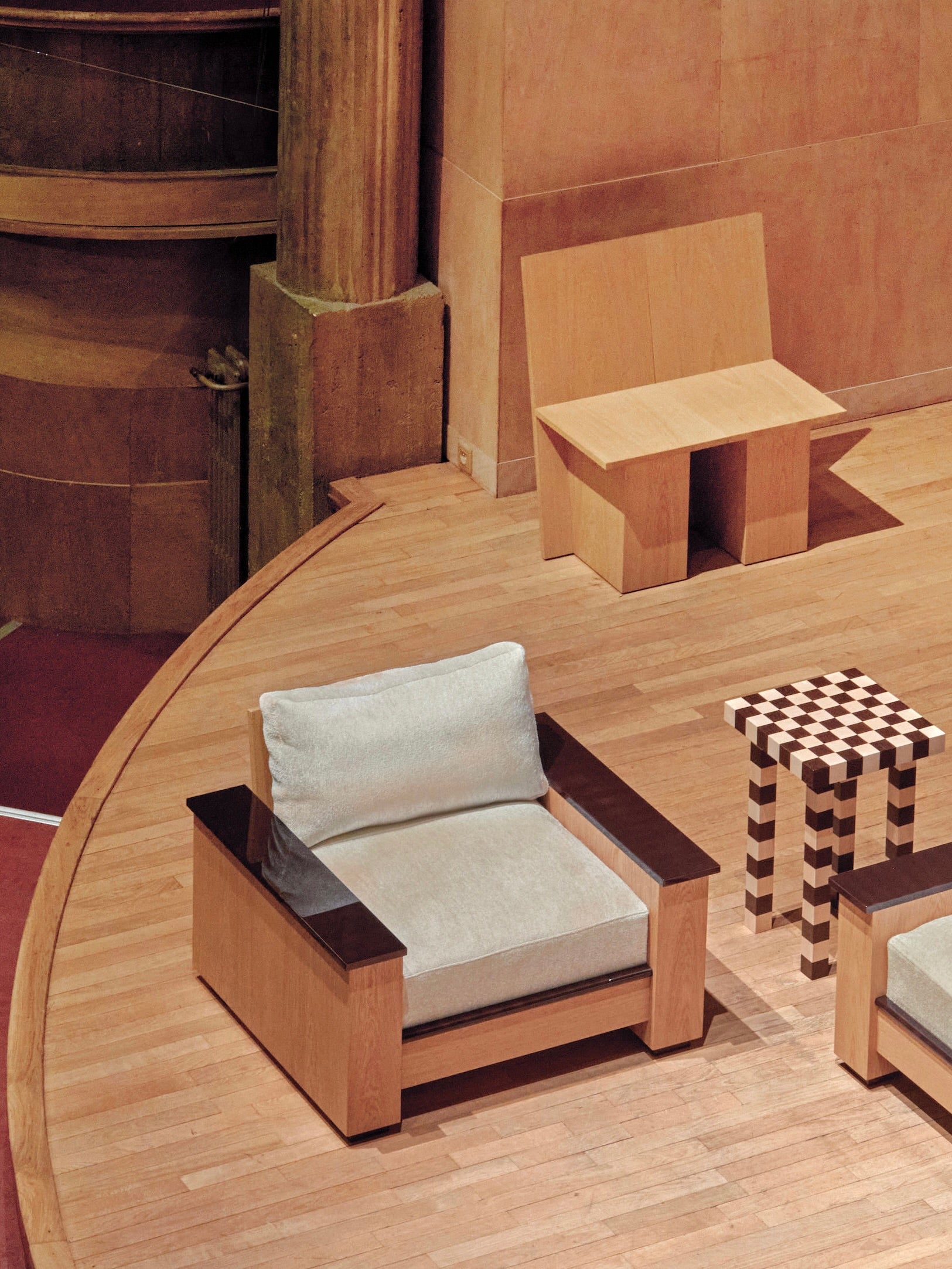 On a wooden stage, two modern chairs are showcased—one is the "Le Capitaine" Arm Chair by HAUVETTE & MADANI, featuring a cushioned light gray seat, while the other is simpler and angular. Between them stands a checkered-patterned table. The chair at the back boasts a unique wooden shell design. The setting is completed by a textured wall and pillar in the background.