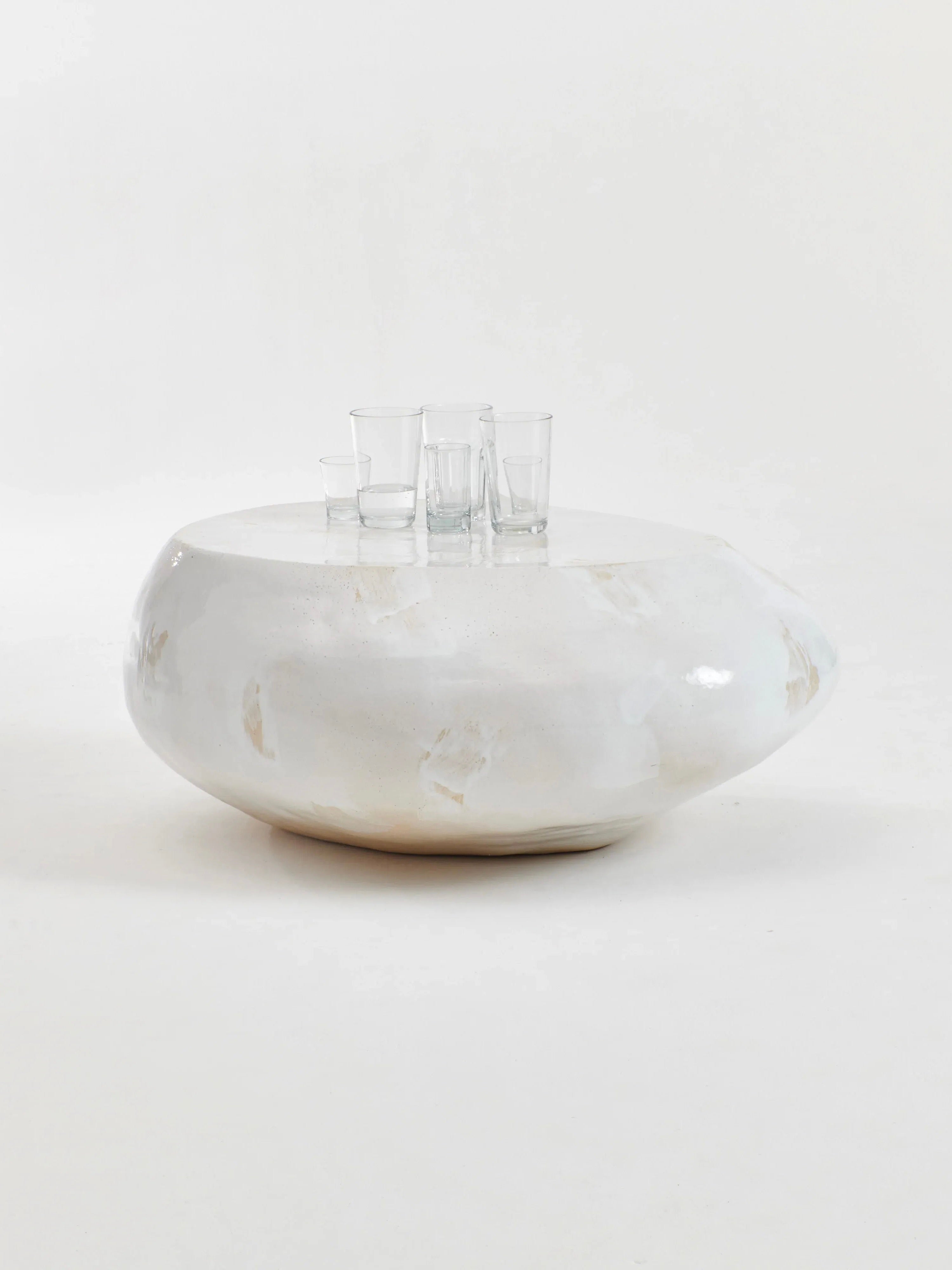 A round, smooth Ceramic Coffee Table in Cream by Project 213A with a glossy finish stands against a plain white background. Four clear glass cups are placed on top of the table. The handmade construction and customized glaze emphasize the minimalist design and simplicity of the monochromatic setting.