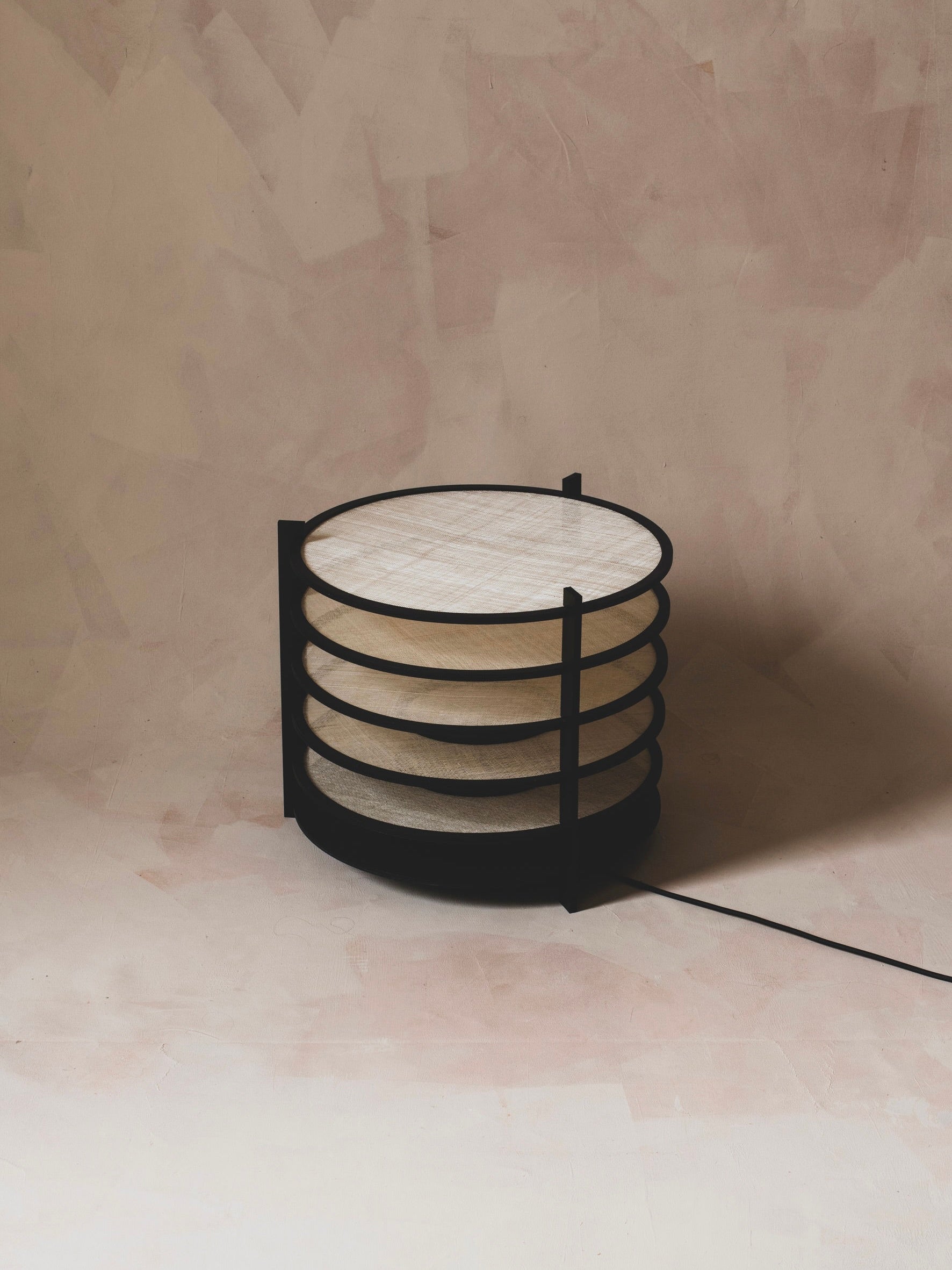 The ELIURPI Sculptural Lamp, featuring a modern circular design with a black metal frame and light wooden shelves, stands against a textured beige background. A slender black cord transforms it into a sculptural piece that infuses any space with artistic ambiance.