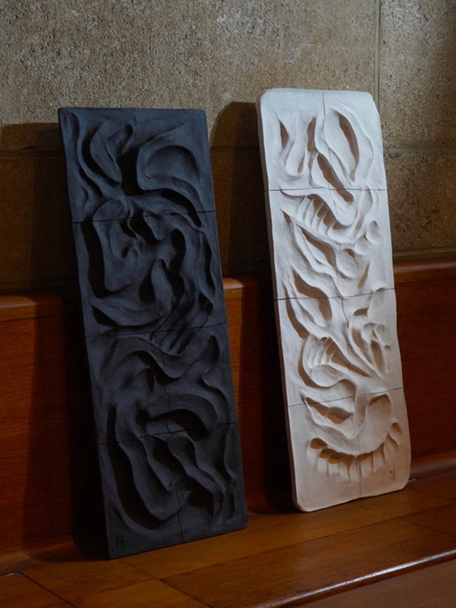 The Kilauea Lava wall sculptures, by Franck Scala, rest against a wooden bench. The black piece features intricate swirls, and the white one has sharp edges. Both sculptures create a striking light-and-dark contrast before a textured wall.