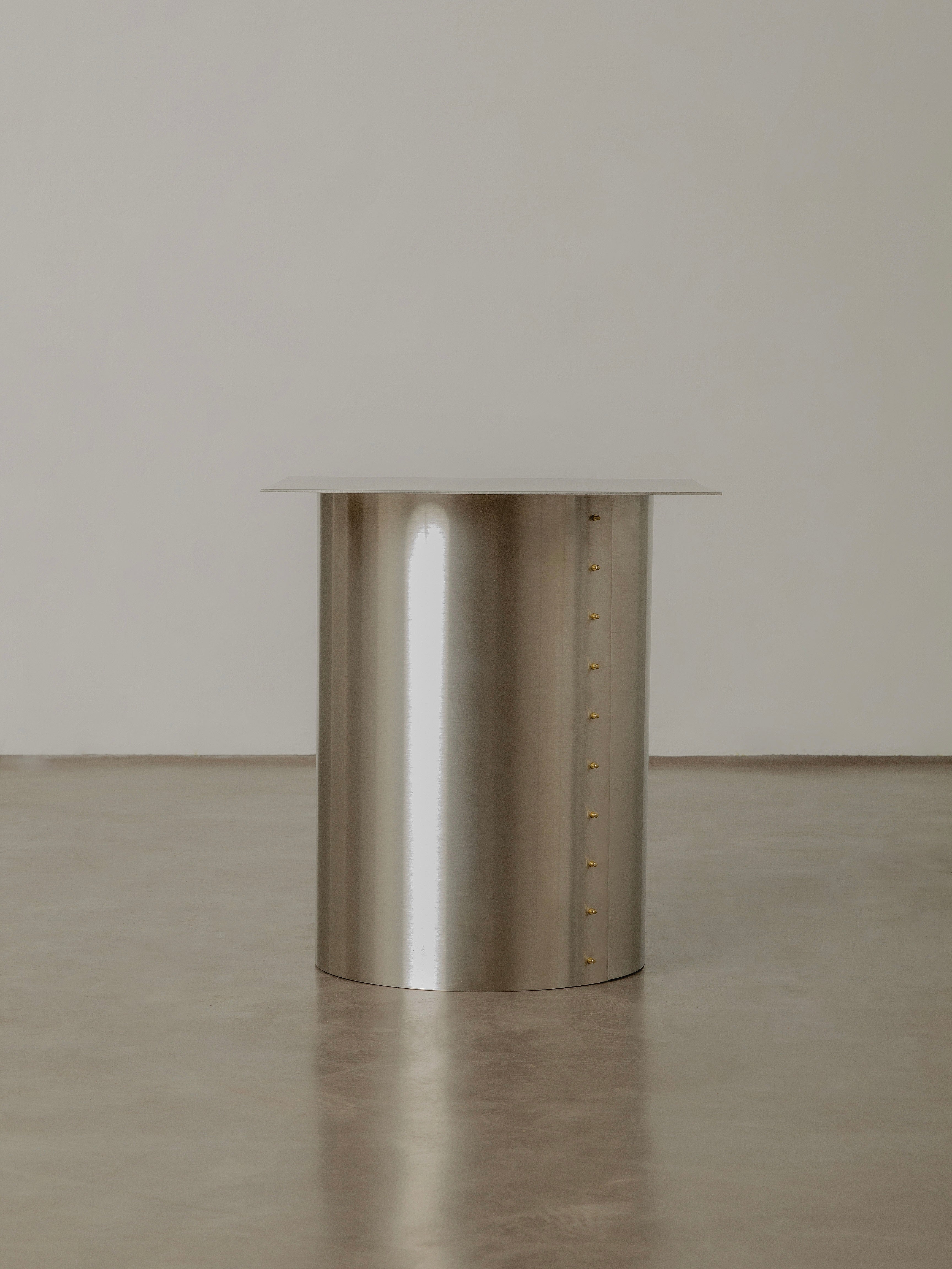 The "Plataforma" Side Table by Pedro Avila, a modern metallic cylindrical piece featuring a smooth reflective surface and rivet detailing, stands on a polished light gray floor against a plain light gray wall, reminiscent of the sleek design of astronomical instruments from the technological era.