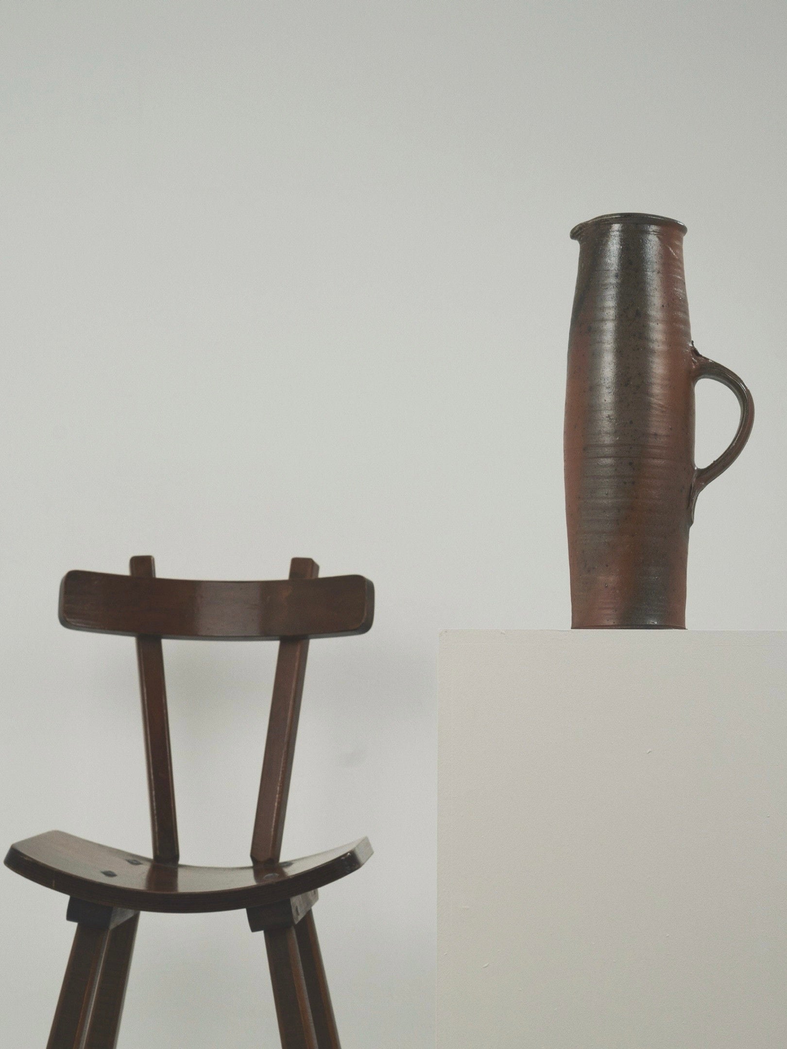 A dark wooden chair sits beside a white pedestal displaying the Large Pitcher by Gustave Tiffoche from Figuresdesign. The tall brown stoneware pitcher with a handle stands out against the plain white background, highlighting its sleek minimalism and French elegance.