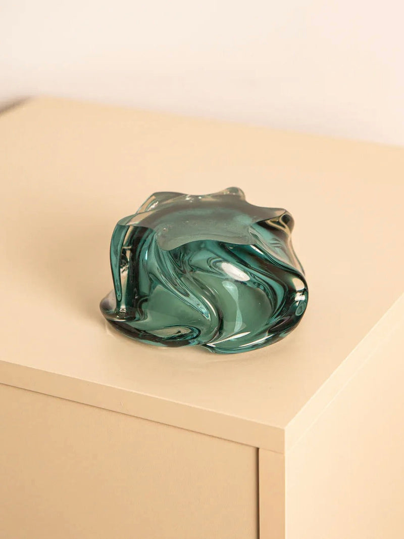 A Dark Turquoise Murano Glass Vide-poches by Treaptyque rests elegantly on a beige tabletop. Its fluid, organic shapes and glossy finish evoke the essence of the turquoise sea, creating a striking contrast against the minimalist Italian design surface.