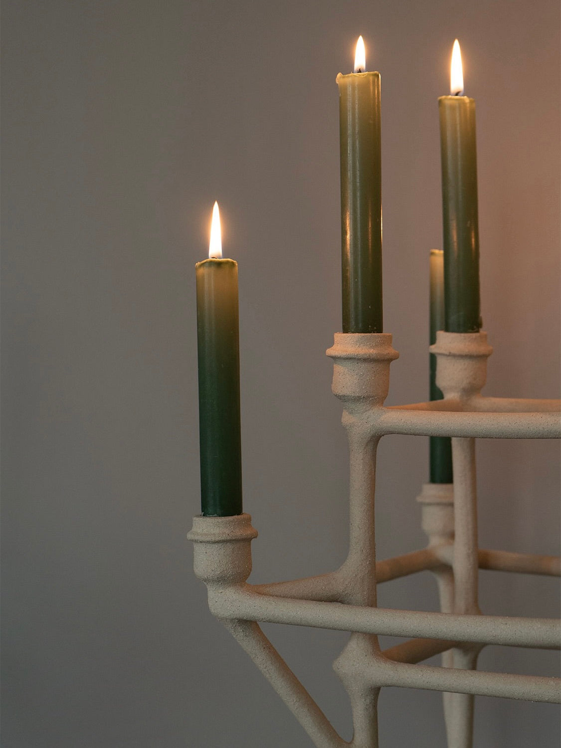 A handcrafted Solenne Belloir Clay Chandelier adorned with green candles, three of which are lit, stands elegantly against a simple gray background.