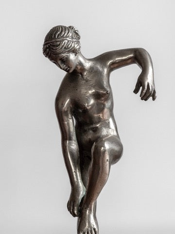 The Elegant Metal Sculpture, 1930s by Spigel features a bronze statue of a graceful, semi-nude woman with an intricate hairstyle. She is bent over with one arm resting on her raised leg and the other arm extended downward, evoking neoclassical design against a plain, light gray background.