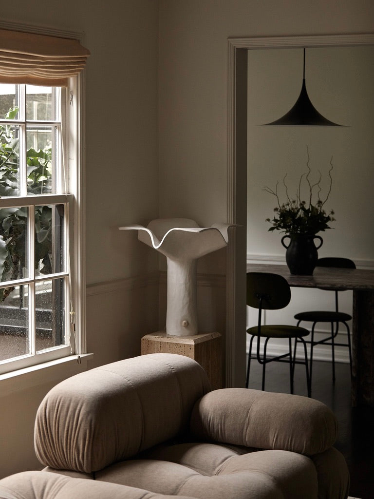 A cozy living space features a plush beige armchair by a window with wooden blinds. The 'La Fleur' Table Lamp by Studio Tristan Kallas elegantly complements a unique white sculpture on a pedestal. A round dining table with chairs and a vase of branches is visible through the doorway.
