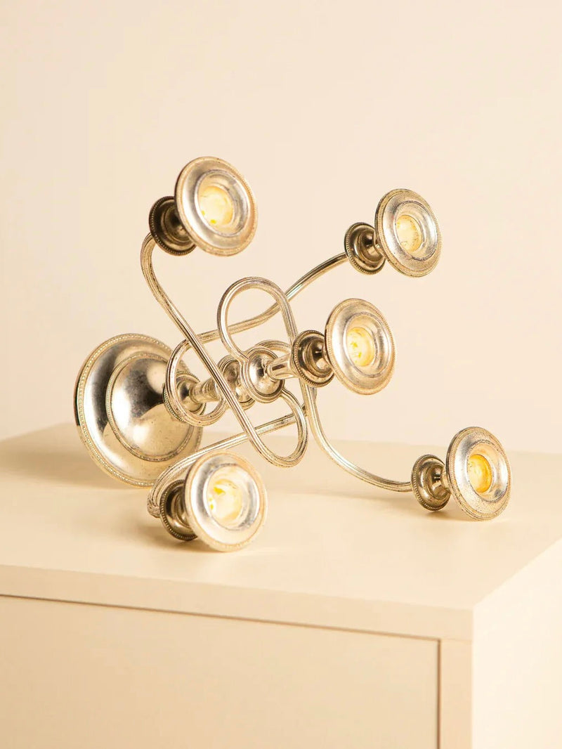 A contemporary abstract metal sculpture, echoing the Treaptyque "Medusa" 60's Italian Candlestick, showcases intertwining loops that support six small circular lights. Displayed on a beige surface against a neutral background, it radiates originality and versatility with a gentle homage to retro aesthetics.