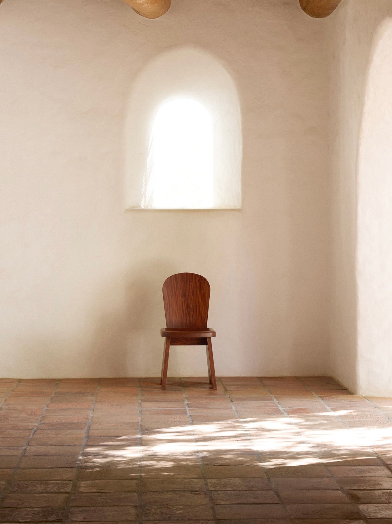 The Rambling Chair in African Mahogany Wood by Lemon exudes contemporary elegance in a minimalist, sunlit room with a white arched window set in a stucco wall. The terracotta tile floor adds warmth, while soft shadows dance from the light streaming through the window.