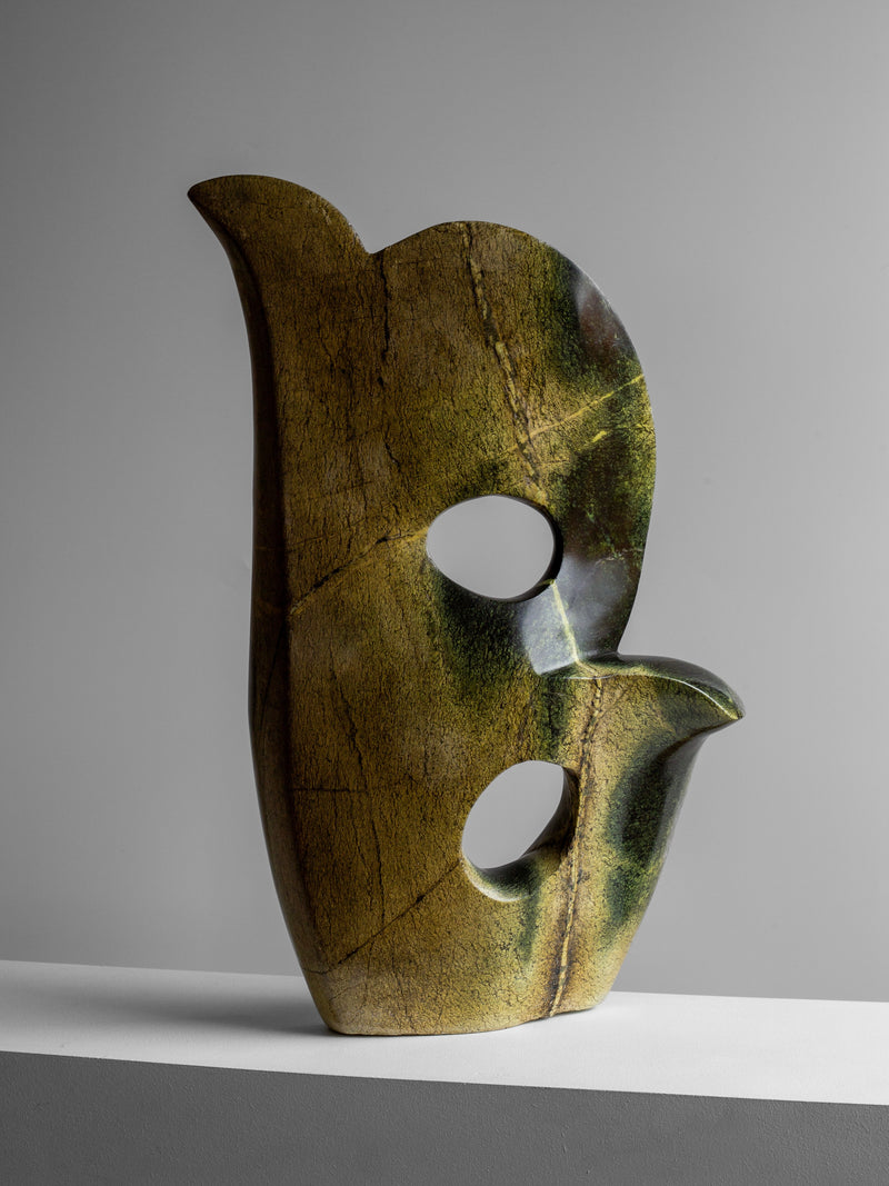 The Spigel "Abstract Stone Sculpture II" by a Zimbabwean artist features a smooth, upward-curving form made from leopard stone. Its green and brown marbled texture comes with two circular cutouts, displayed elegantly against a neutral gray background on a white surface.