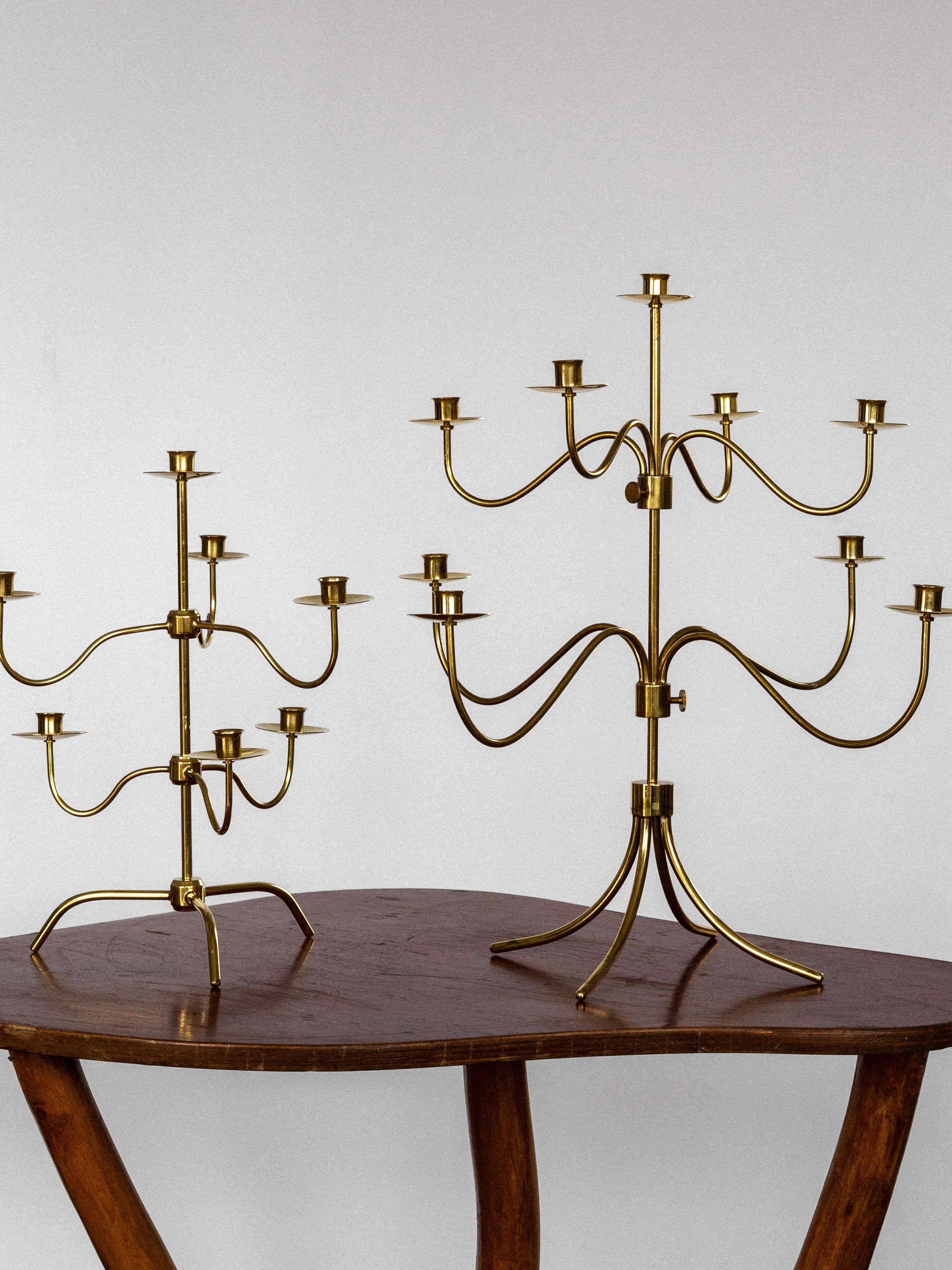 Two Medium Mid-Century Brass Candelabras by Spigel are displayed on a wooden table with curved legs against a plain background. The candelabra on the left is smaller with simpler design features, while the one on the right is larger and has more elaborate branching arms.