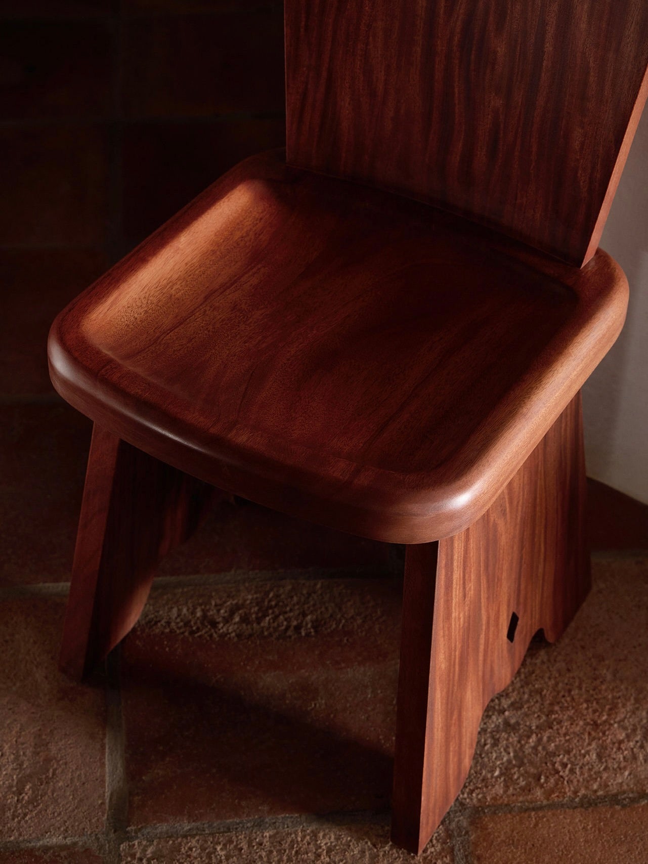 The Rambling Chair from Lemon, crafted in African Mahogany wood, showcases a smooth, polished seat and backrest with rich wood grain. Set on warm-toned tiled floors, the lighting accentuates its contemporary elegance and sleek design.