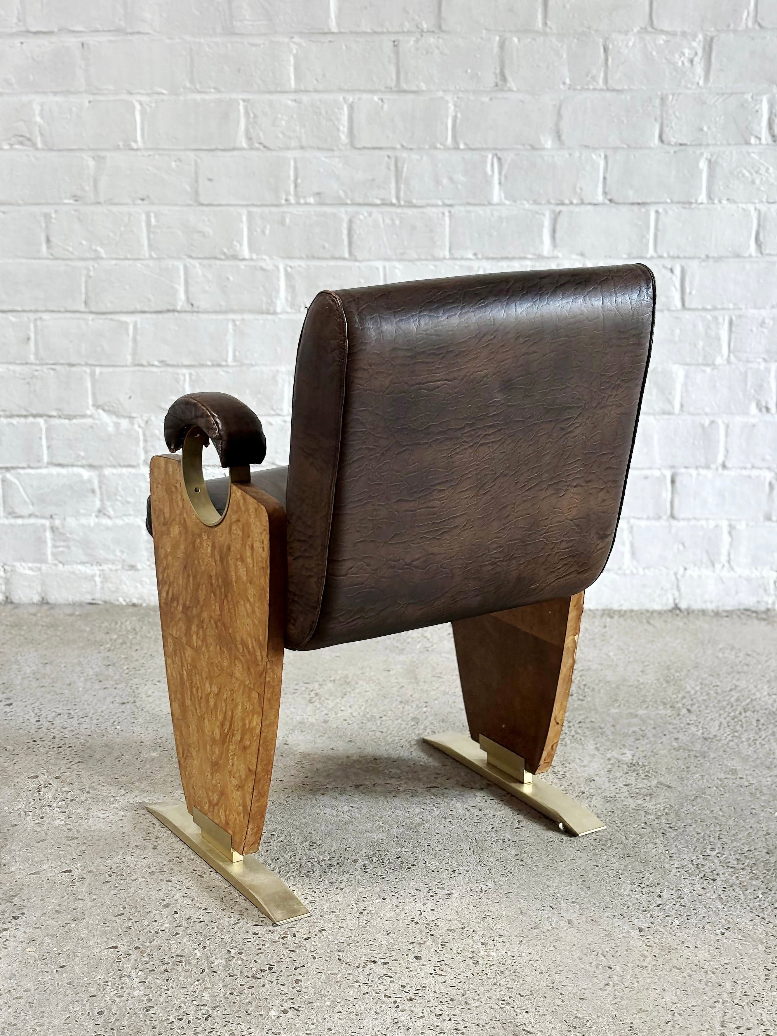 Introverso's Art-Deco Style Side Chair from the 1970s boasts a brown skai leather backrest and armrests, burl walnut sides, and sleek gold metal legs, perfectly positioned on a concrete floor against a white brick wall.