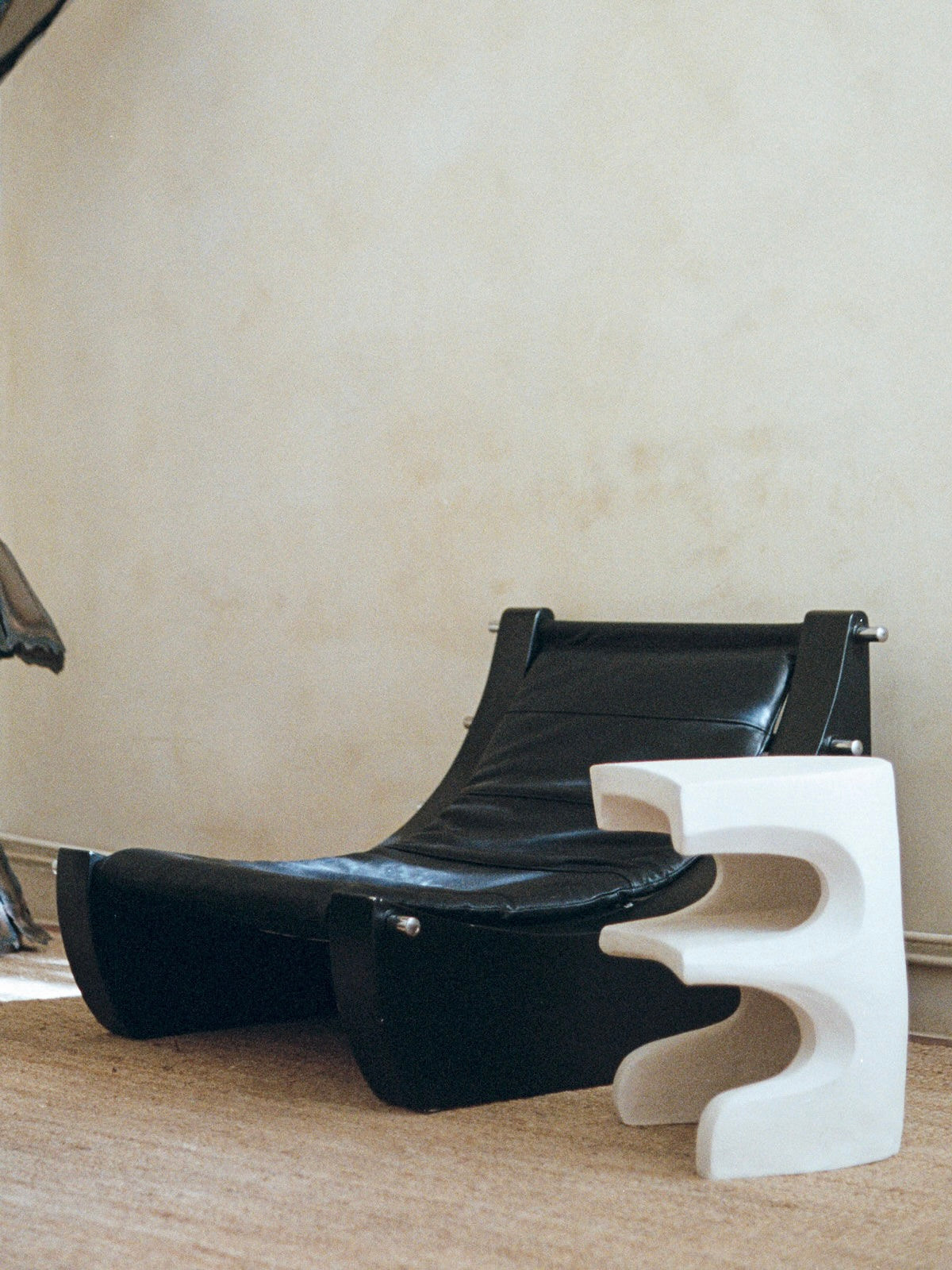 A modern black leather lounge chair rests on a beige carpet beside the Baisers Volés - Carved Stoneware Coffee Table by Lea Bigot, featuring sculptural curved cut-out designs, with a textured light-colored wall in the background.