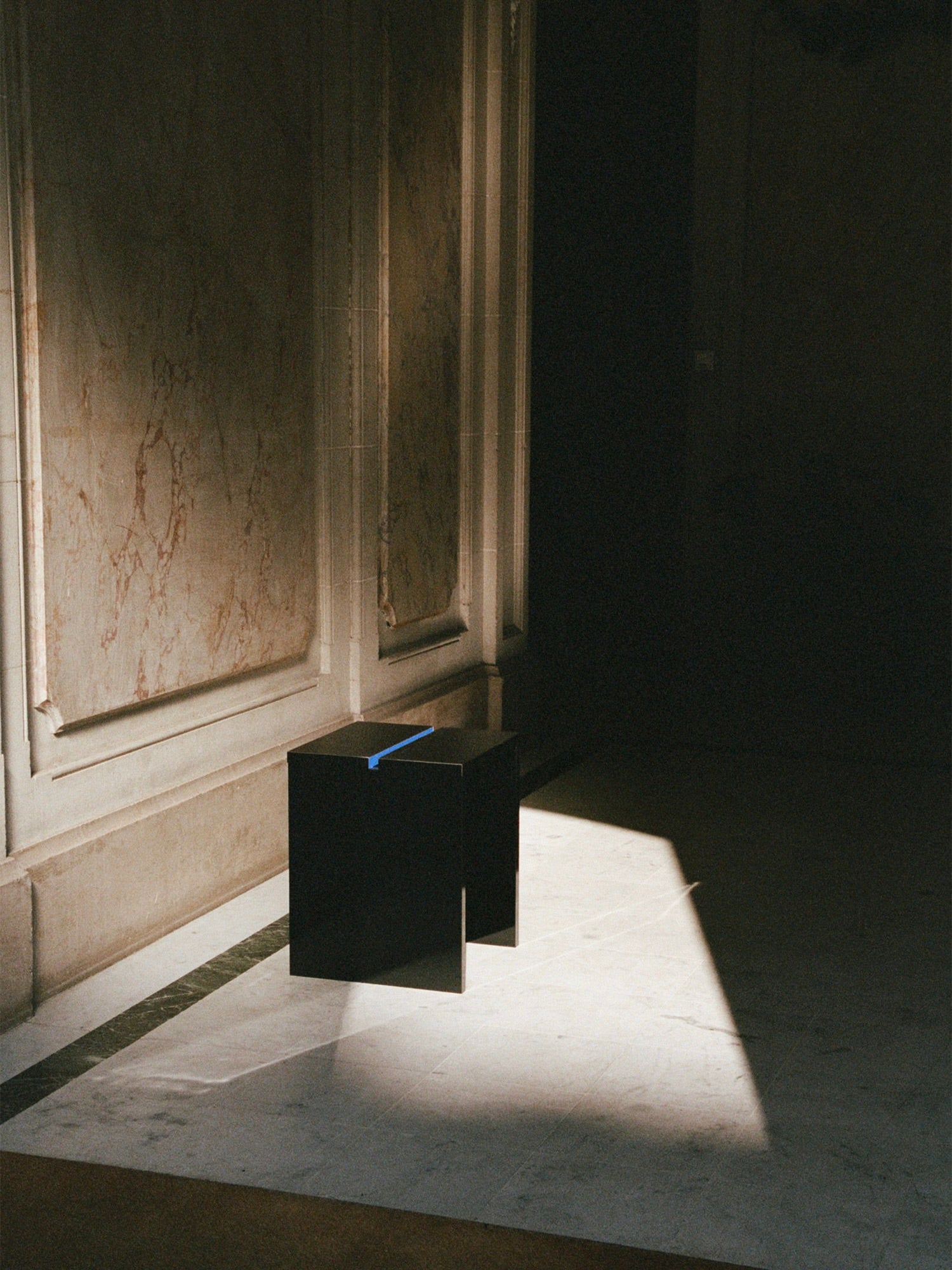 A TEGET Mara Gap Stool – Wet Black, cube-shaped, basks in sunlight on a marble floor, casting a shadow. Positioned by a beige marbled wall, it contrasts the bright shaft of light with the dim surrounding space.