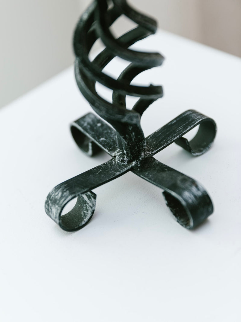 Close-up of the Bottega Jacobs Forged Candleholder – Brutalist Design 1950s, a hand-forged, black, spiral-shaped metal piece with a looped base on a white surface. The design features four curled legs supporting a twisted vertical structure, embodying the Brutalist style.