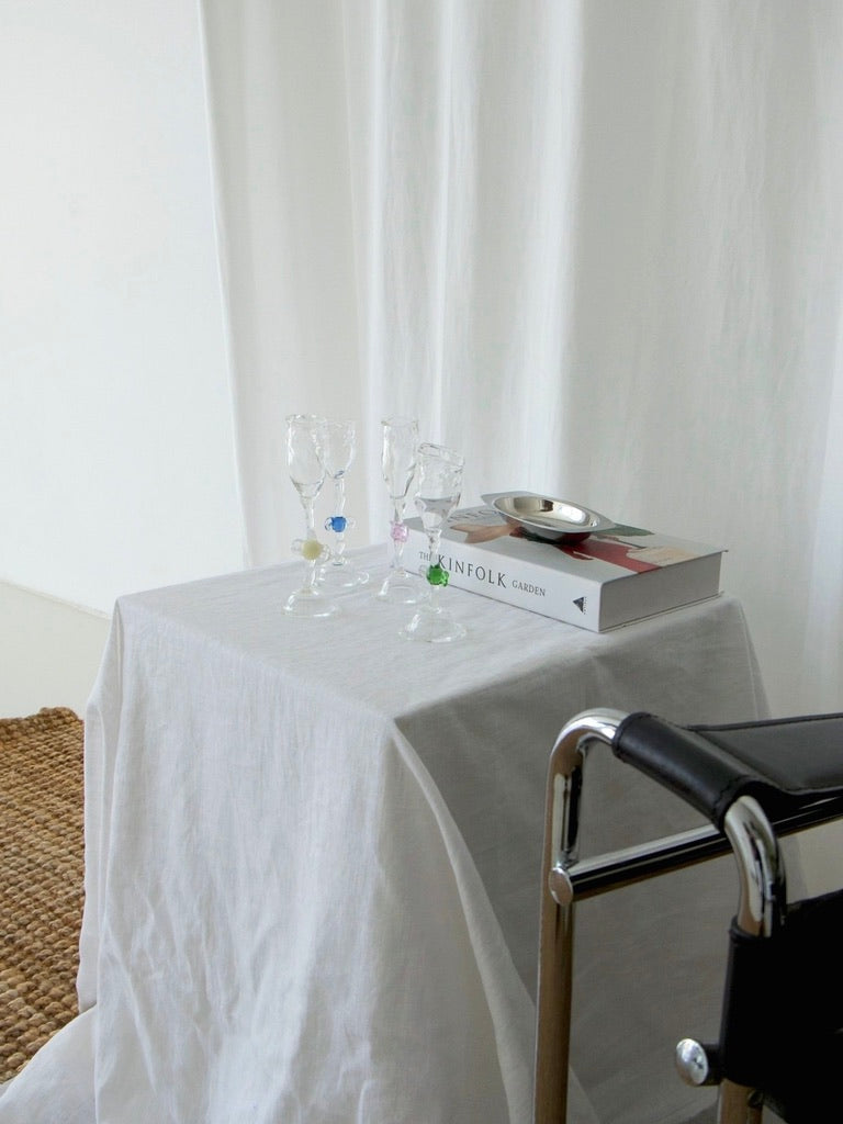 A small table with a white cloth displays two Vivian Alarcón Mouth Blown 'Candy' Wine Glasses, a stack of books, and a silver dish. A black metal-framed chair is nearby, with white curtains in the background.