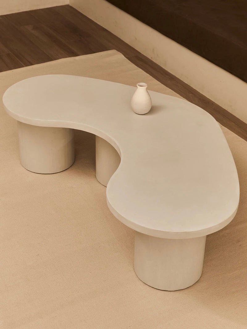 The "Parmigiano" Coffee Table by MarlotBaus, with its minimalist kidney-shaped design and smooth curves, elegantly rests on a light rug. A small white vase adorns its center, enhancing the sophisticated ambiance of the room with wooden floors and brown cushioned seating in the background.