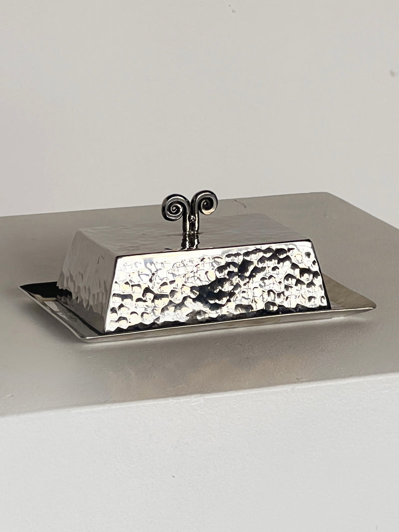 The Spiral Butter Dish by Porter Studio, with a hammered rectangular design and decorative handle, sits on a light surface. The lid exudes 1970s charm, featuring a handle with two outward-facing swirls.