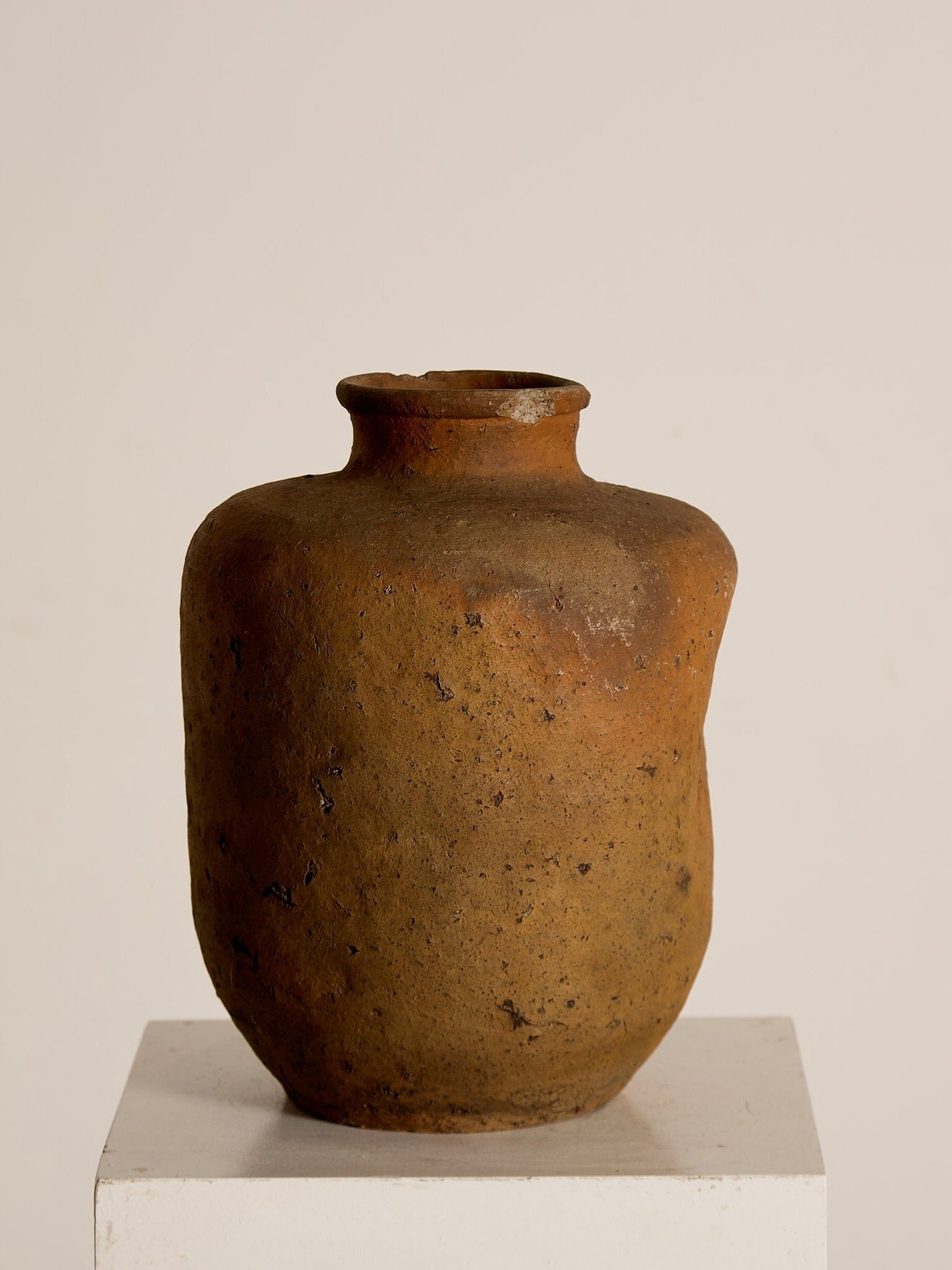 The Japanese Stoneware Vessel by Ancien et Jolie features a rustic, speckled glaze and irregular surface. This brown ceramic vase with a narrow neck stands elegantly on a white pedestal, creating texture against the plain background.