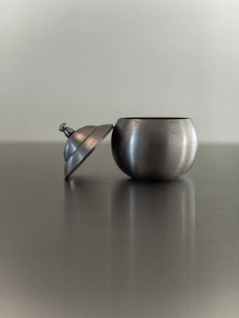 A compact, circular Steel Bonbonnière by Out For Lunch, featuring a matching lid that's slightly tilted and set on a reflective surface. The soft, blurred background enhances the industrial charm and elegance of its design.