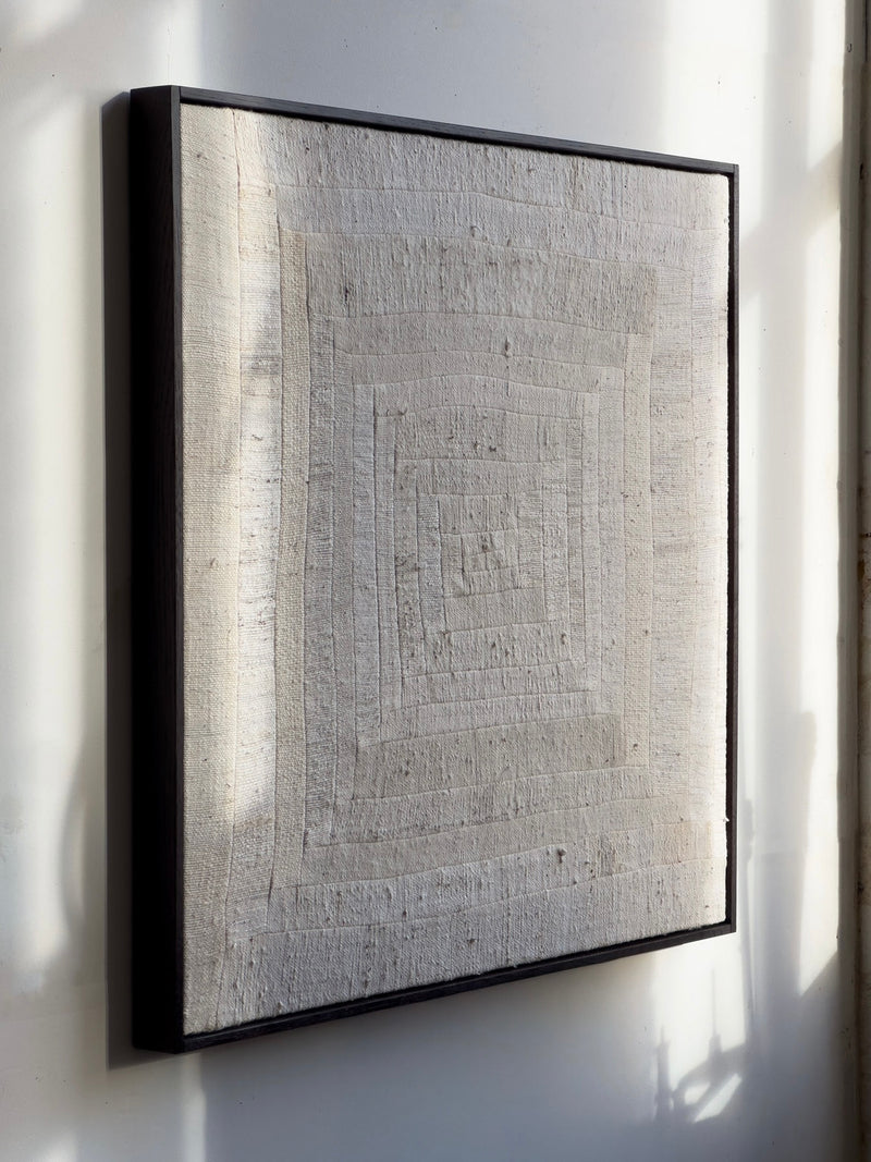 Bec Kirby’s 'To Hold', 2025, is a textured off-white square canvas inspired by log cabin patchwork with concentric raised rectangles for a subtle 3D effect. Framed in black solid oak, it creates an intriguing shadow on the white wall when lit naturally.
