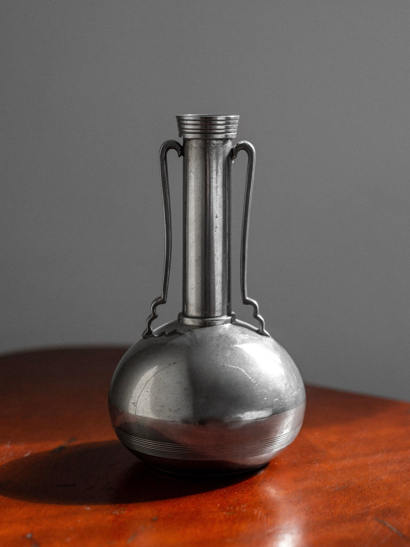 An Art Deco Metal Vase, 1935 by Spigel with a shiny, metallic finish, narrow neck, decorative handles on either side, and a rounded base is placed on a wooden surface. The background is a plain gray wall. Light casts subtle reflections on the vase, highlighting its Scandinavian craftsmanship from GAB (Guldsmedsaktiebolaget).