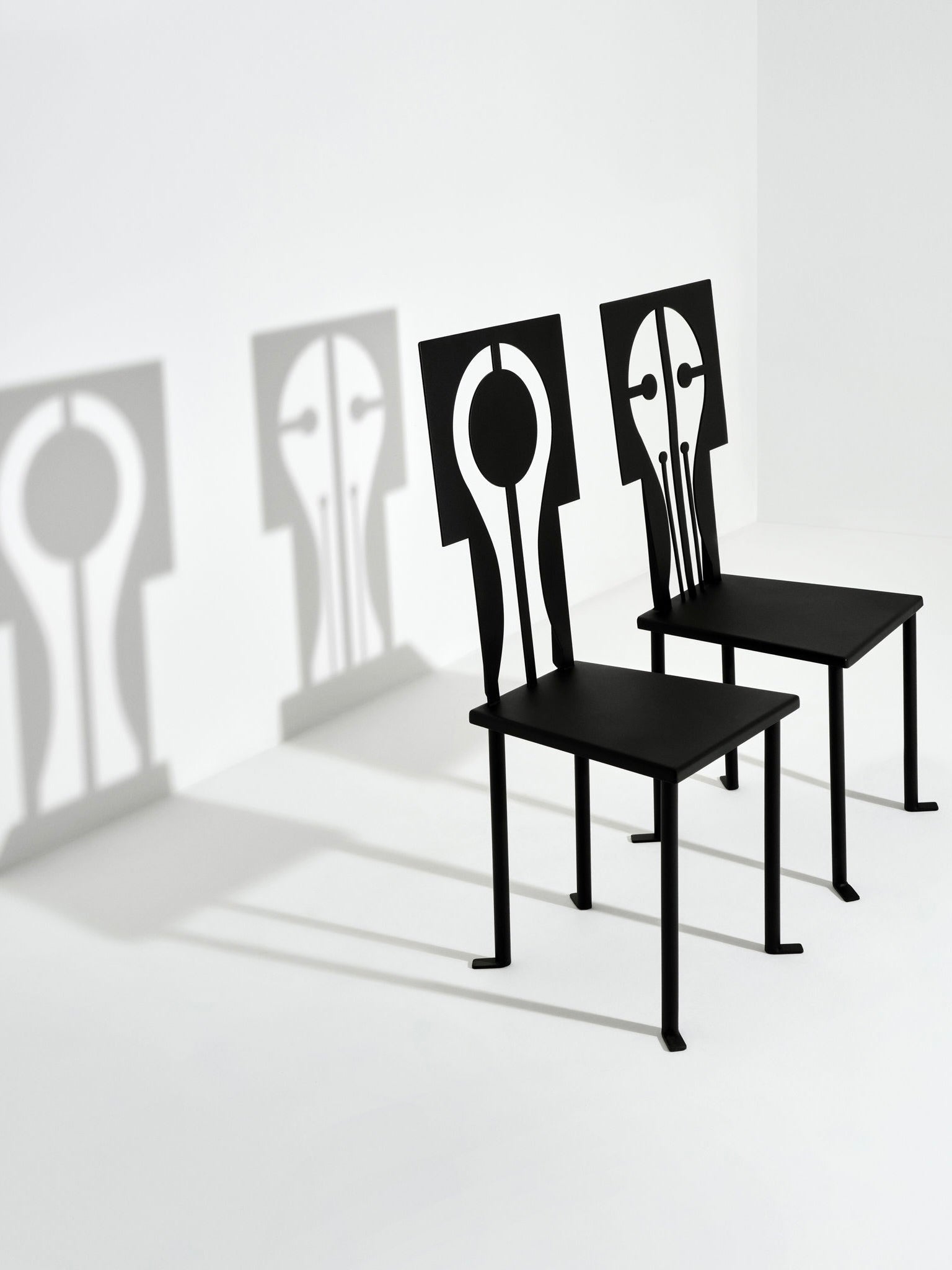 Two Luna Chairs in Iron and Black Powder Coat by Animate Objects showcase artistic cutout backs, casting abstract shadows resembling faces on a plain white wall. The design of these handcrafted pieces creates unique and interesting patterns with the light.