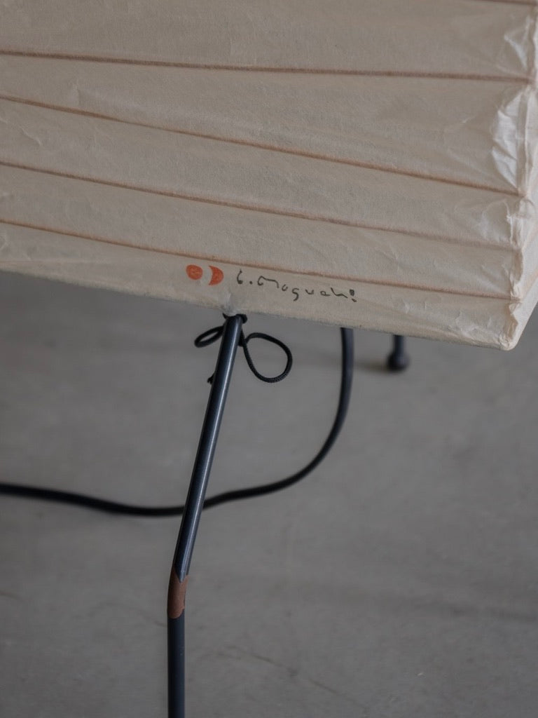 A close-up of the Bicci de' Medici Isamu Noguchi - Akari UF lamp showcases a white pleated paper shade and slender black metal legs, echoing mid-century modern aesthetics. A small orange logo and text on the shade reflect Noguchi's iconic style against a plain background.